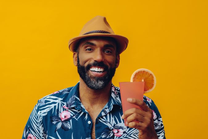Happy male in hat and colorful shirt holding cocktail with yellow background