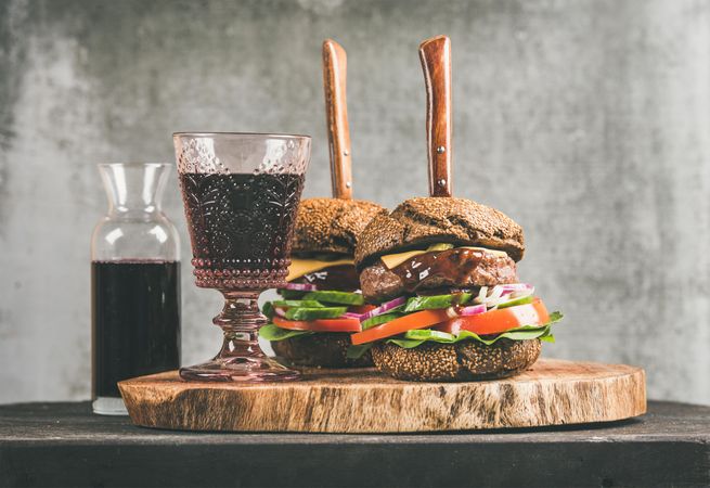 Two cheeseburgers, stacked with fresh vegetables, on wooden board, knifes, with wine, and decanter