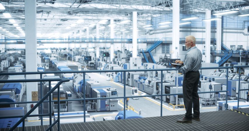 Experienced Male Engineer Standing on a Platform, Using Laptop Computer and Overlooking Production at a Modern Automated Electronics Manufacture with AI Support. Factory Executive at Work