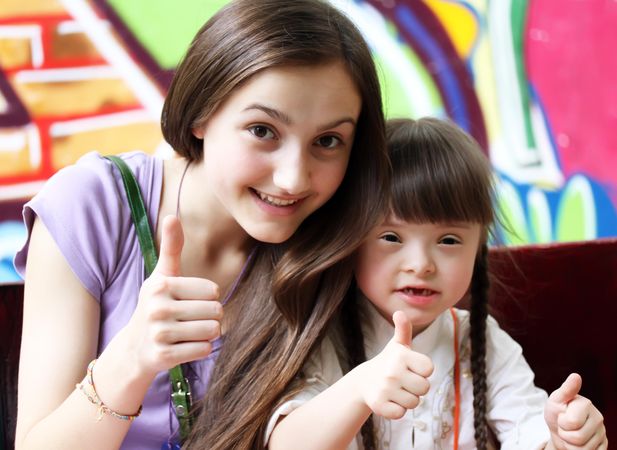 A teenage girl and her younger sister with Down syndrome doing a thumbs up sign