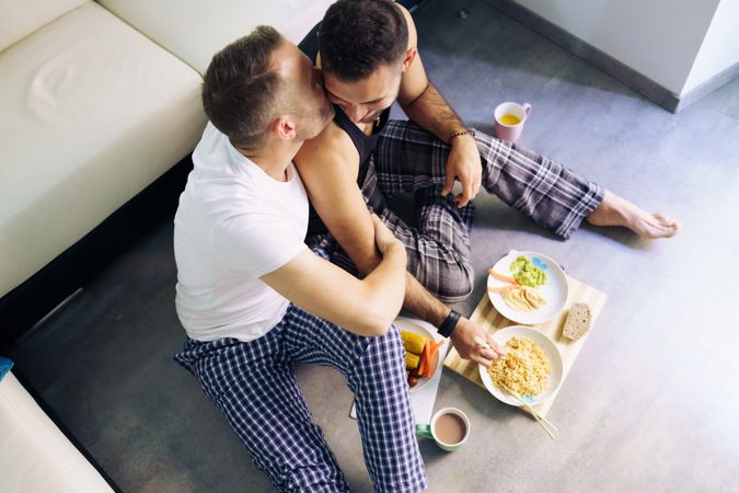 Looking down at male couple embracing while eating meal together at home sitting on floor