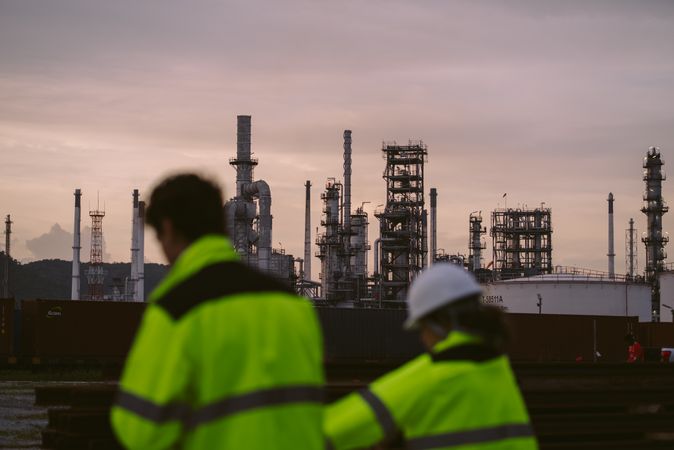 Two industrial workers, wearing high-visibility jackets, focus on background of refinery factory