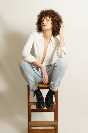 Self-assured woman in jeans and boots sitting on wooden stool in studio shot