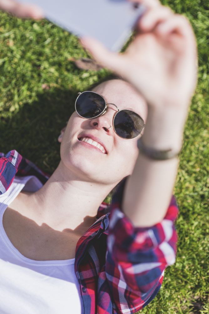 Above view of a young smiling hipster woman lying on grass in a sunny day at a park while taking a selfie with a mobile phone