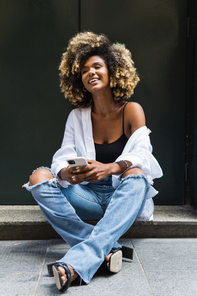 Smiling Black woman using smartphone while sitting on concrete border