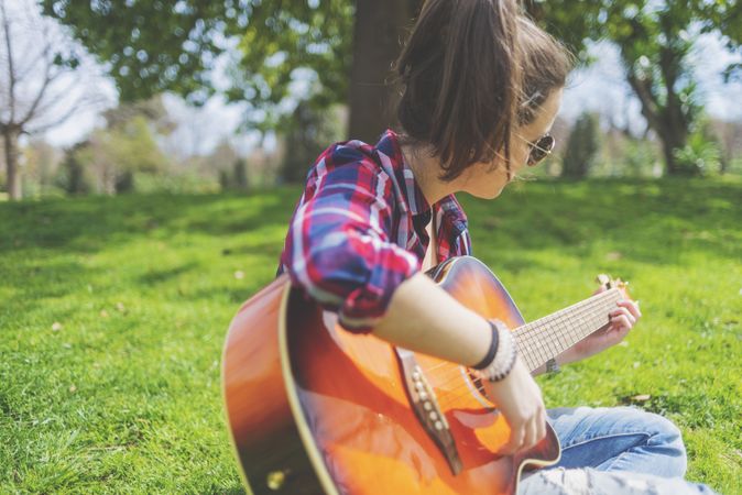 Front view of a young hipster woman wearing sunglasses, sitting on grass in a park while enjoying playing guitar