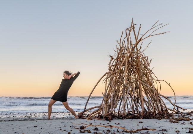Young woman dancing on shore near water and wooden twigs placed for campfire