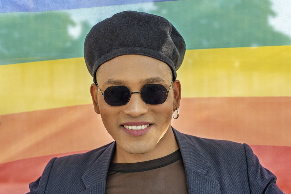 Young Latino gay man with make-up on wearing a fashionable hat and sunglasses, isolated on a Rainbow flag background. LGBT.