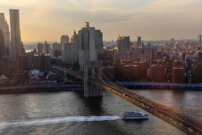 Iconic New York City Landscape Over East River with Skyscrapers, Brooklyn Bridge, Cars and Ferry Boats. Cinematic Evening Urban Skyline with Sunset and Lightly Clouded Sky