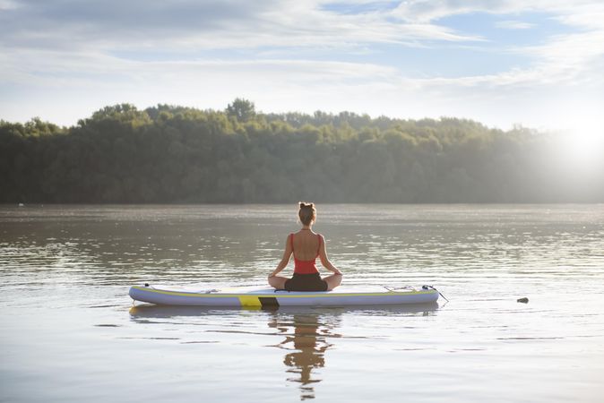 Woman meditating on paddleboard on fresh lake in the morning surrounded by trees