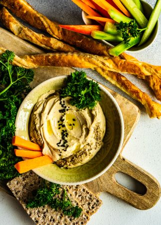 Top view of creamy hummus dip in bowl on board served with veggies, bread sticks and rye crackers