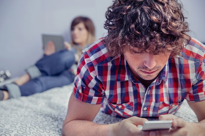 Man using smartphone over bed and woman in background reading