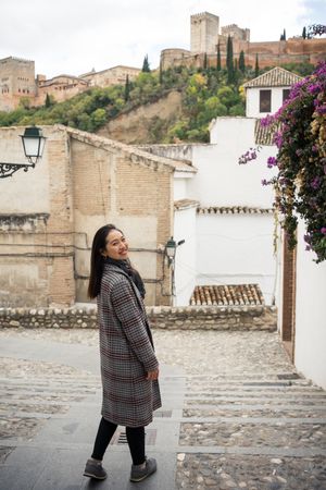 Asian female traveler in warm wear smiling among old buildings near big castle