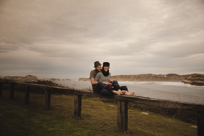 Young guy in hat near elegant woman playing on hand drum in cap on seat near coast of sea and cloudy sky