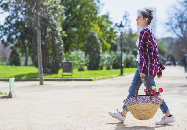 Side view of a young hipster woman holding a wicker basket while walking in a park in sunny day