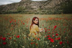 adult woman looking over shoulder at camera while sitting in field with poppies and daisies