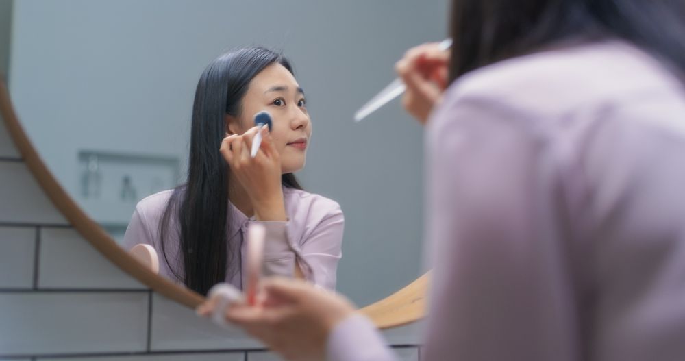 Stylish South Korean Female Using a Soft Brush to Put Tonal Foundation Cream in a Bathroom at Home.