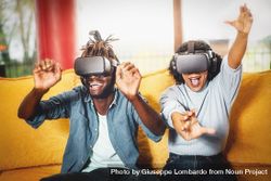 Couple of happy friends enjoying a VR game wearing goggles sitting on a yellow sofa bE9pl6