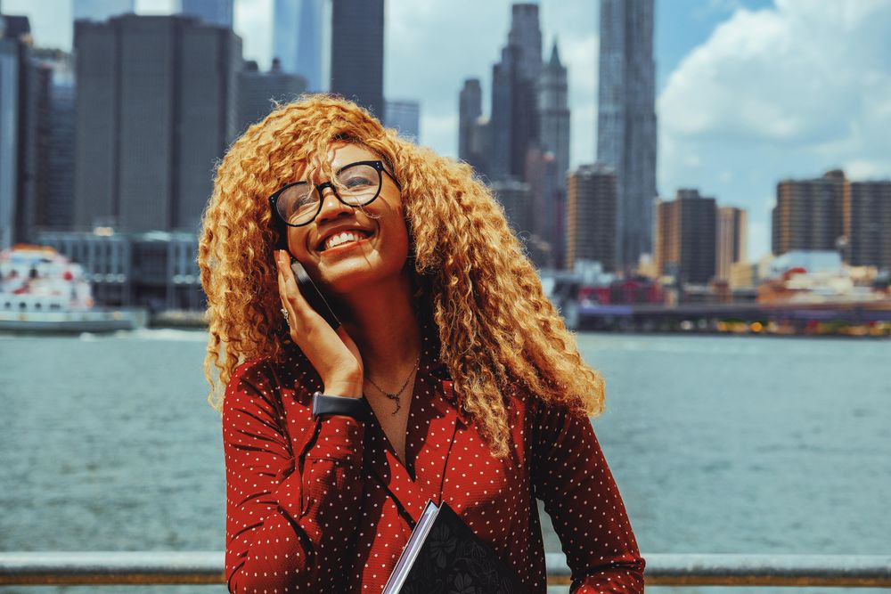 Smiling Black female speaking on smart phone by Hudson River