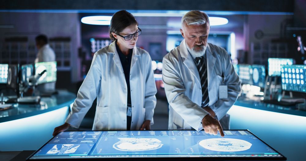 Female Neuroscientist And Male Surgeon Talking, Using Interactive Touch Screen Table With CT Scan Of Human Brain With Tumor, Finding Treatment