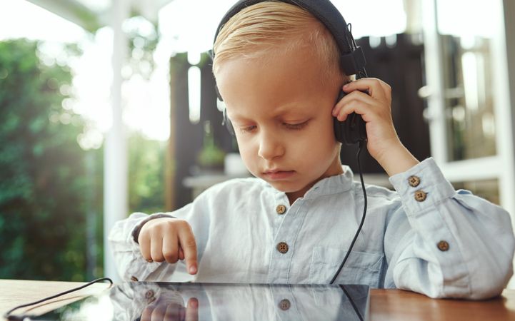Serious blond boy using headphones and selecting show or game on digital tablet