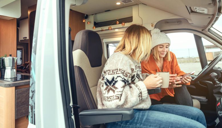 Side view of female friends sitting in parked van having coffee and checking social media on phone