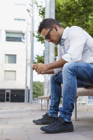 Side view of male sitting on bench looking down at phone