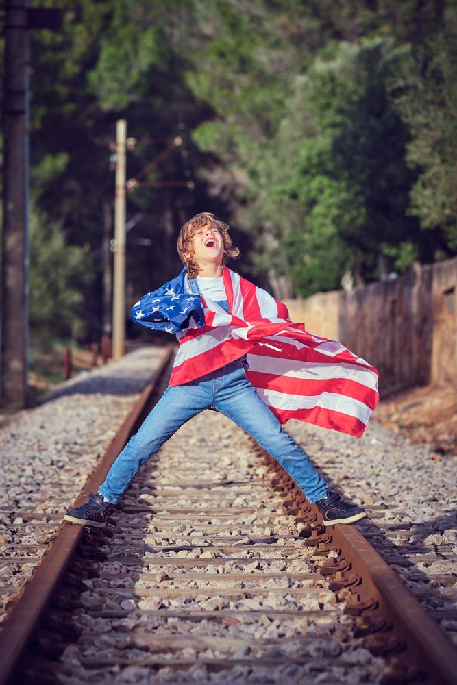 Joyful preteen boy wrapped in American flag standing on rails