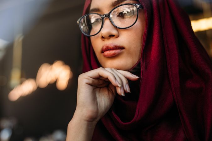 Close up portrait of young woman wearing hijab and eyeglasses with her hand on chin looking away