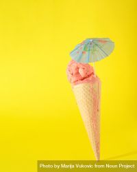 Cone with pink scoop of ice cream with cocktail parasol on yellow background 0gKJ30