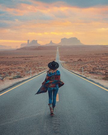 Back view of woman wearing bohemian style jacket and hat walking on the road in the middle of the desert
