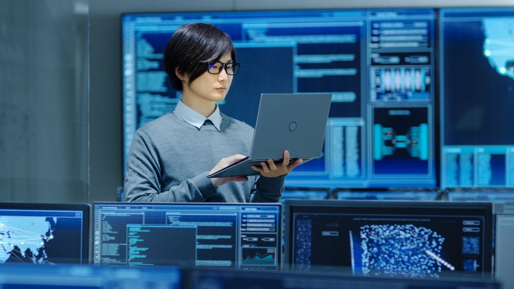 In the System Control Room IT Technician Holds and Works on a Laptop, in Background Multiple Displays with Graphics.