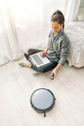 Woman freelancer sitting on floor and switching robotic vacuum