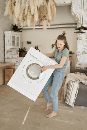 Joyful strong woman carrying washing machine in kitchen