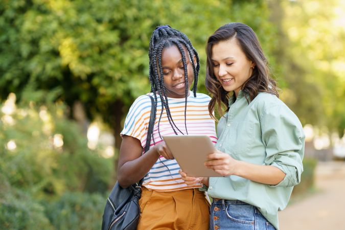 Female friends pointing at something on tablet in casual clothing in park