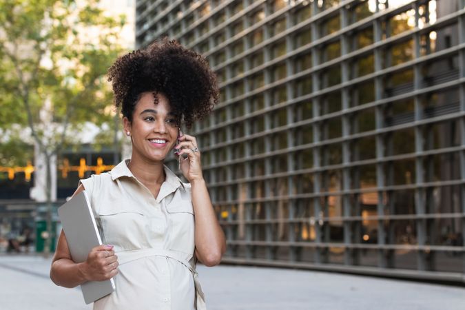 Smiling Black pregnant woman talking on smartphone and carrying a laptop