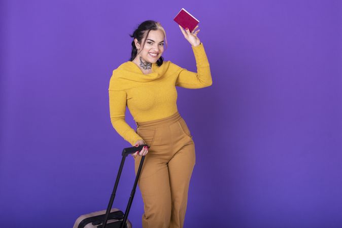 Young happy woman traveler with suitcase while showing her passport against purple background. Copy space.