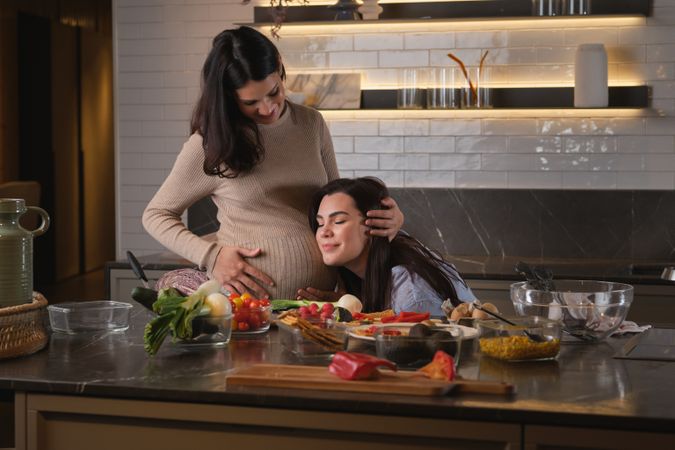 Woman putting head on belly of pregnant woman in kitchen