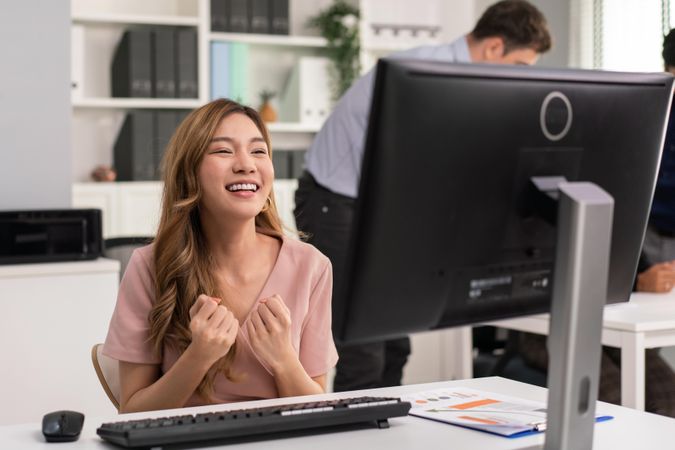 Asian woman making fists in celebration while sitting at her desk
