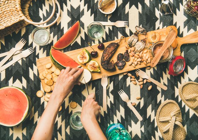 Woman cutting cheese on board at summer picnic on pueblo pattern blanket, with snacks