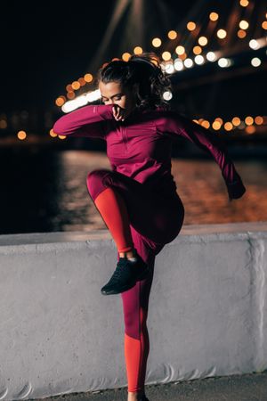 Woman in dark red sportswear doing jumps near lit bridge in evening