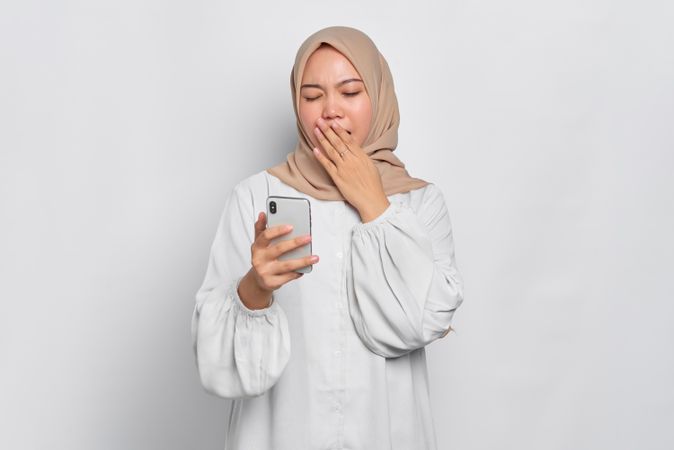 Tired Asian female in headscarf yawning while using her cell phone