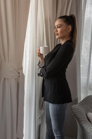 Side view of woman in dark blazer drinking coffee standing by the window