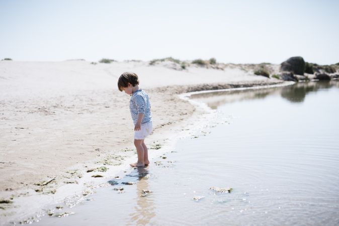 Barefoot child in short watching waves on sandy beach