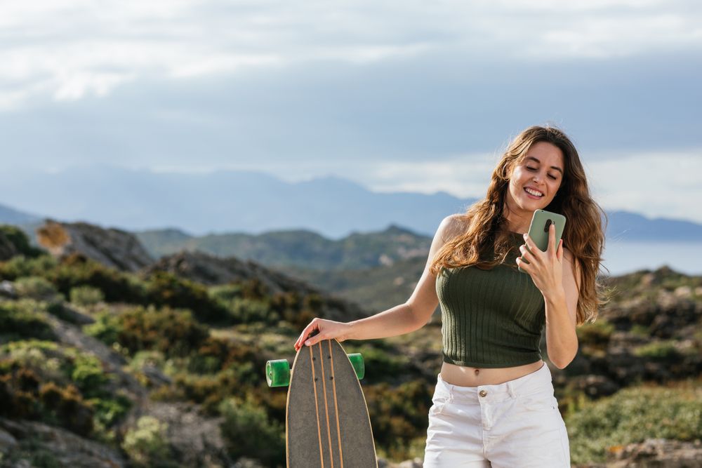 Cheerful skater chatting on smartphone against ridges in summer