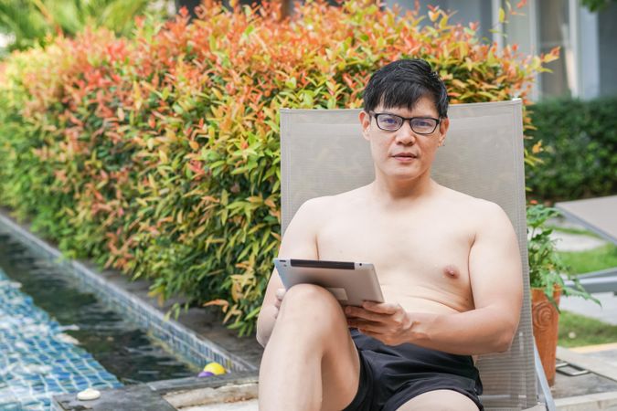 Man sitting by pool reading on digital tablet