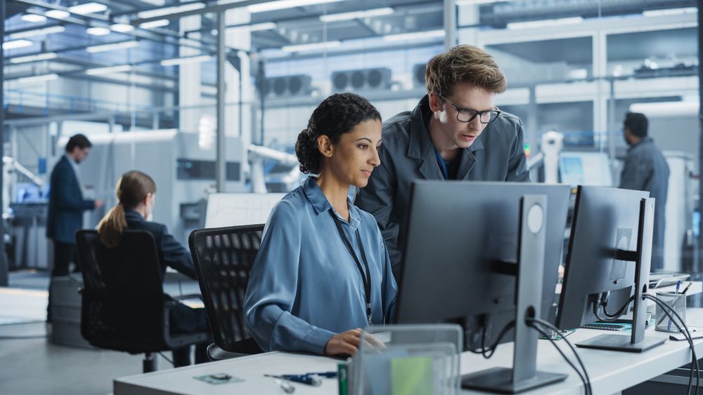 Portrait of a Young Male Assembly Line Worker and Hispanic Female Engineer Collaborating on a Project, Using Computer and Talking in a Factory with Robotic Arms Producing Devices for Tech Industries.