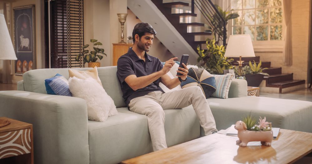 Cheerful Young Indian Man Comfortably Using His Smartphone At Home