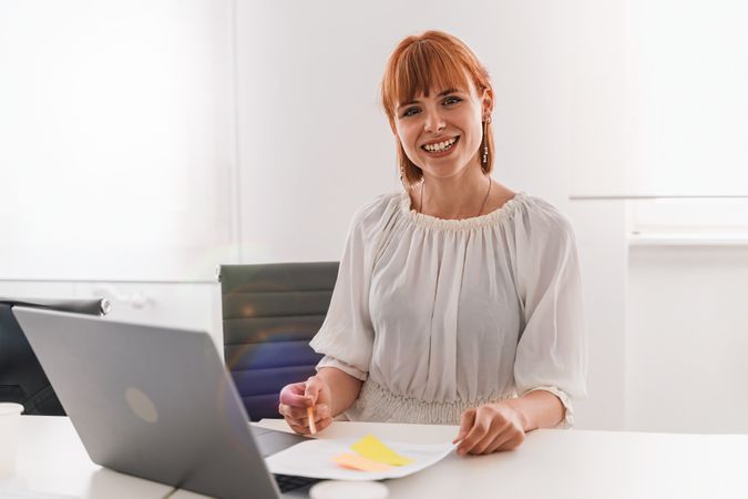 Redhead woman with nose piercing smiling at camera, sitting with pen, laptop, papers, and sticky notes in bright office