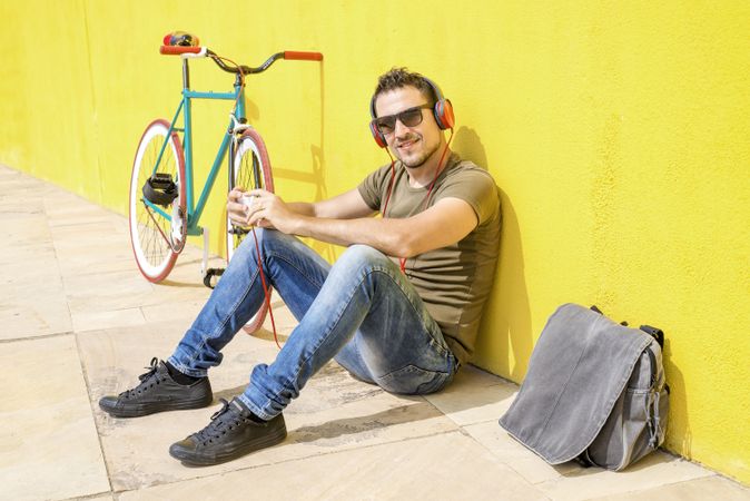 Happy male looking at camera in front of yellow wall next to bike and listening to music
